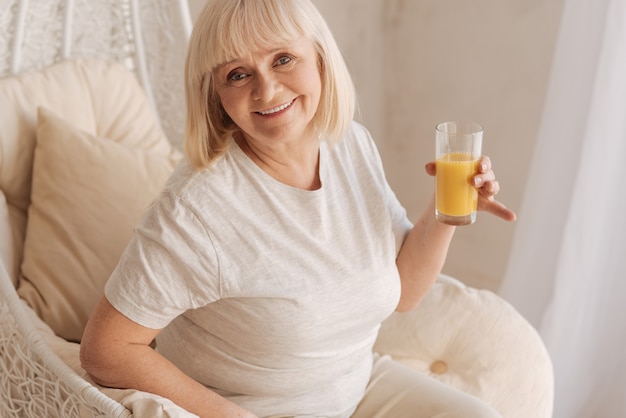 Cheio de vitaminas. Mulher enérgica e saudável alegre segurando um copo com suco de laranja e sorrindo enquanto está de bom humor