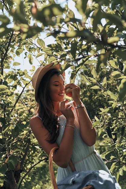 Cheira bem! Mulher jovem e atraente cheirando uma maçã e sorrindo em pé no jardim