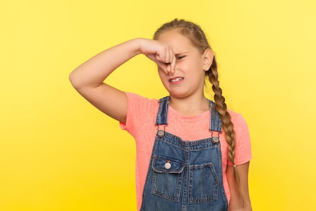 Cheiro horrível. Retrato de menina com trança em macacão jeans prendendo a respiração, apertando o nariz e expressando nojo ao fedor, odor intolerável. tiro de estúdio interno isolado em fundo amarelo