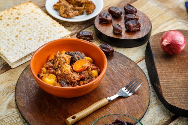 Chelnt prato judeu com carne na mesa em um prato em um suporte ao lado dos ingredientes e pão ázimo.
