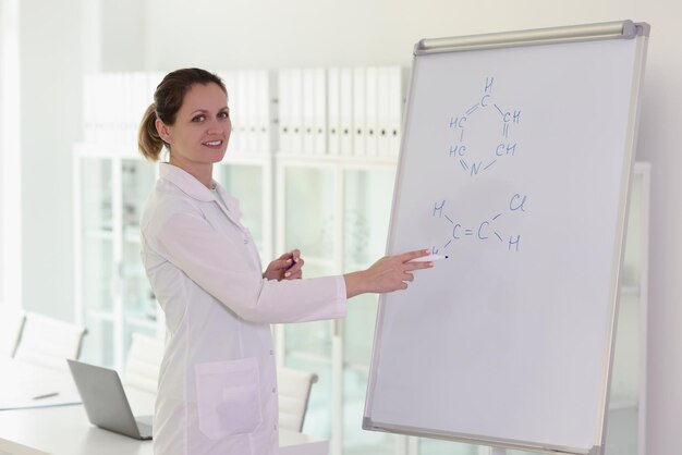 Chemikerin schreibt Formeln auf großes Whiteboard, lächelt und schaut in die Kamera, die Frau dirigiert