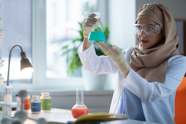 Chemisches Glas. Dunkeläugiger Chemiker mit Kopftuch, der chemisches Glas mit blauer Flüssigkeit hält