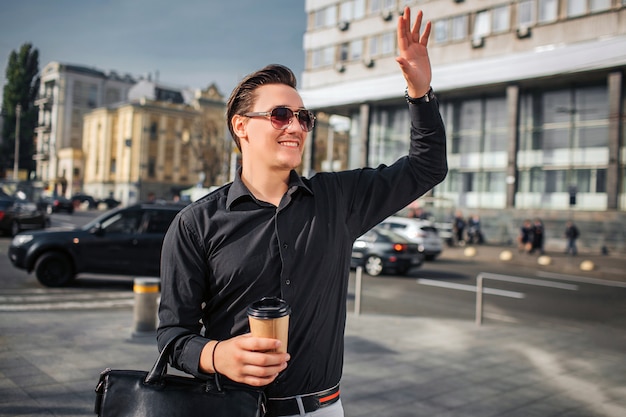 Chherful y feliz joven camina en la calle. Él saluda con la mano. Guy tiene una taza de bebida y una bolsa.