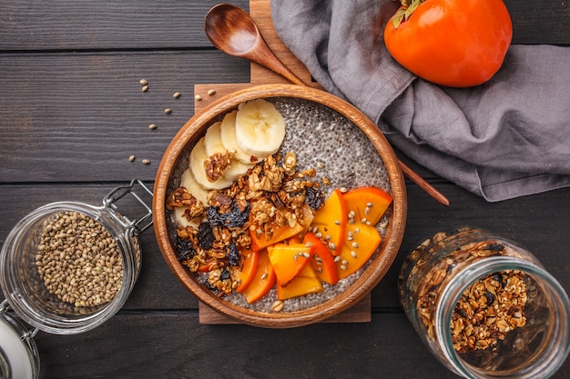 Chia-pudding mit banane, persimone und granola, dunkler hölzerner hintergrund, draufsicht, kopienraum.