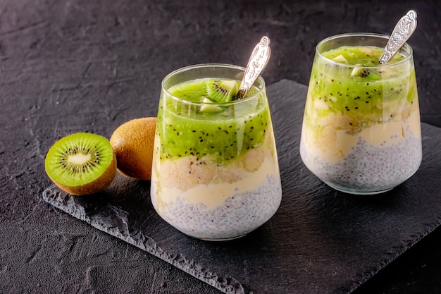 Chia-Samen-Pudding mit Kiwi, Banane und Mango. Gesundheits-Entgiftungsfrühstück in Gläsern auf schwarzem Steinhintergrund.