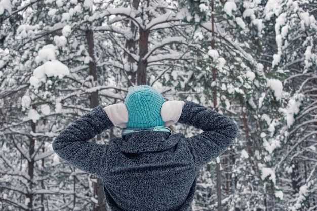 Una chica con un abrigo gris y un sombrero de punto azul claro se encuentra en un hermoso bosque nevado de invierno en un día nublado y nevado