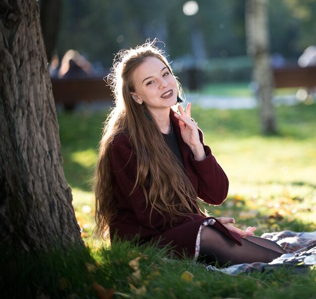 Chica con abrigo sonriendo sentada en un plaid en el parque de otoño