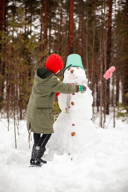 Chica en abrigo verde en invierno en el bosque cerca de muñeco de nieve