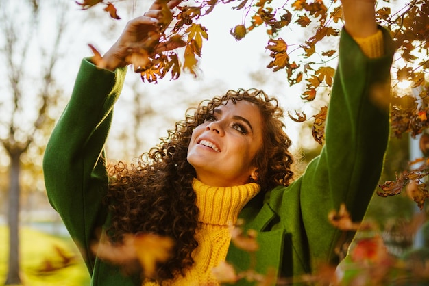 Chica con un abrigo verde y un suéter amarillo bajo el color de las hojas de otoño que rompe el árbol