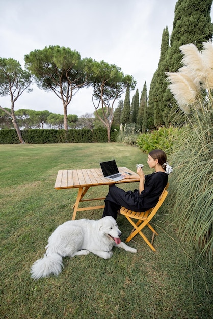 Chica acaricia a perro y usa la computadora portátil en la mesa en el césped