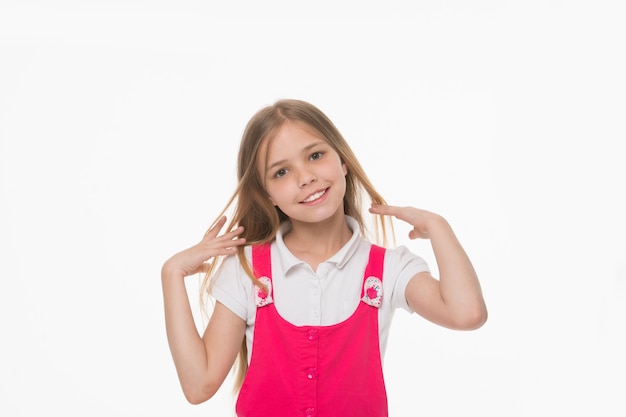 Chica acariciando su largo cabello rubio. Niño encantador con una sonrisa encantadora, la belleza y el concepto de infancia. Niño en traje rosa aislado sobre fondo blanco. Chica juguetona con grandes ojos color avellana posando en estudio.