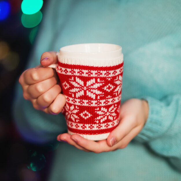 Chica en un acogedor suéter de punto bebiendo café de una taza de punto rojo