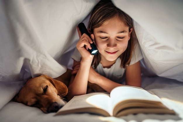 Chica acostada en la cama con su perro debajo de una manta leyendo un libro a altas horas de la noche
