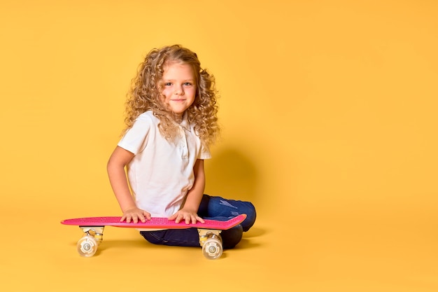 Chica activa y feliz con el pelo rizado, auriculares divirtiéndose con penny board, patineta con soporte de cara sonriente