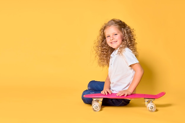 Chica activa y feliz con el pelo rizado, auriculares divirtiéndose con penny board, patineta con soporte de cara sonriente