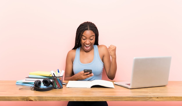 Chica adolescente afroamericana estudiante con cabello largo trenzado en su lugar de trabajo sorprendida y enviando un mensaje