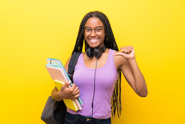 Chica adolescente afroamericana estudiante con largo cabello trenzado sobre pared amarilla aislada orgullosa y satisfecha