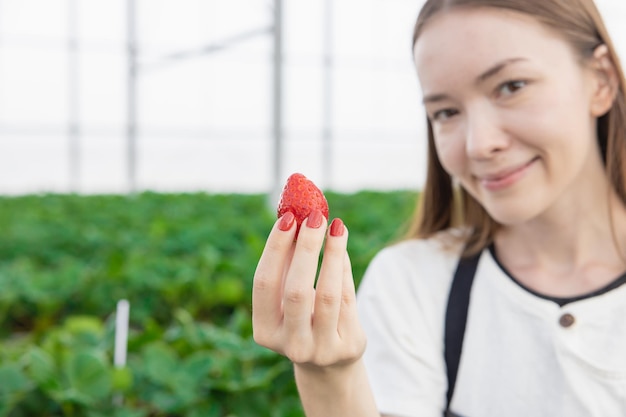 Chica adolescente agricultora que muestra una gran fresa dulce fresca roja de la granja orgánica de la casa verde interior