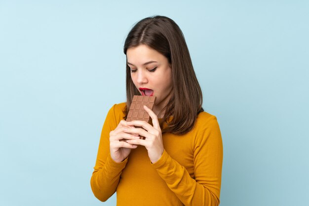 Chica adolescente aislada en la pared azul comiendo una tableta de chocolate