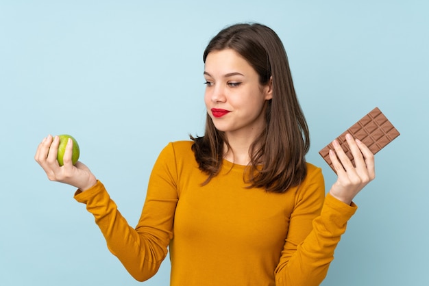 Chica adolescente aislada en la pared azul que tiene dudas mientras toma una tableta de chocolate en una mano y una manzana en la otra