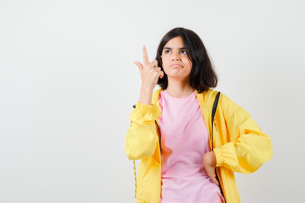 Chica adolescente apuntando hacia arriba en camiseta, chaqueta y mirando confiado, vista frontal.