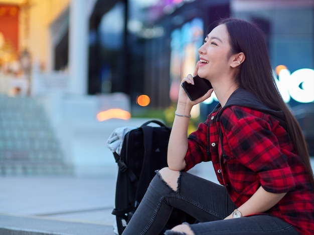 Chica adolescente asiática feliz hablando por teléfono móvil sentado frente al centro comercial