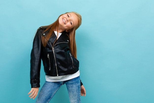 Chica adolescente bastante caucásica con largo cabello castaño en chaqueta negra y jeans sonrisas aisladas sobre fondo azul