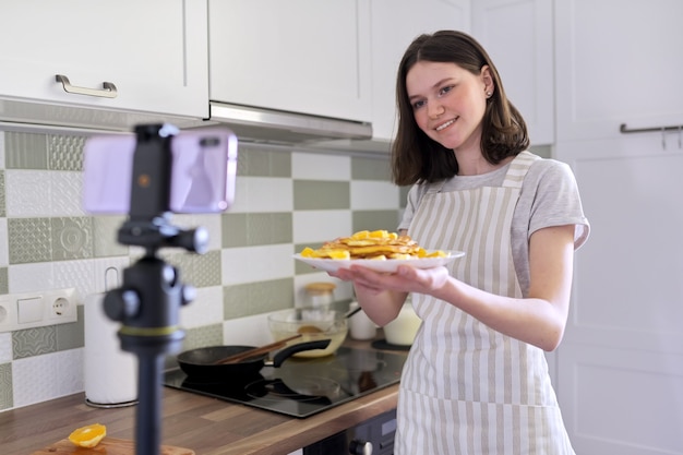 Chica adolescente, blogger de comida cocinando panqueques con naranja en casa en la cocina, filmando receta de video. Mujer con comida preparada, buen provecho. Pasatiempos, canal de videos con seguidores, niños y adolescentes