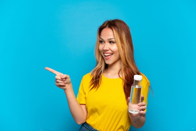 Chica adolescente con una botella de agua sobre fondo azul aislado apuntando con el dedo hacia el lado y presentando un producto