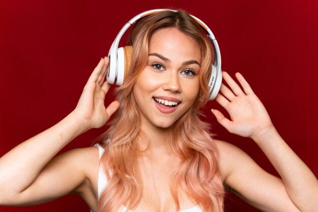 Chica adolescente con cabello rosado escuchando música sobre pared roja aislada