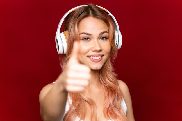 Chica adolescente con cabello rosado en rojo aislado escuchando música y con el pulgar hacia arriba