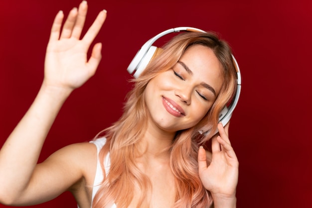 Chica adolescente con cabello rosado sobre pared roja aislada escuchando música y bailando