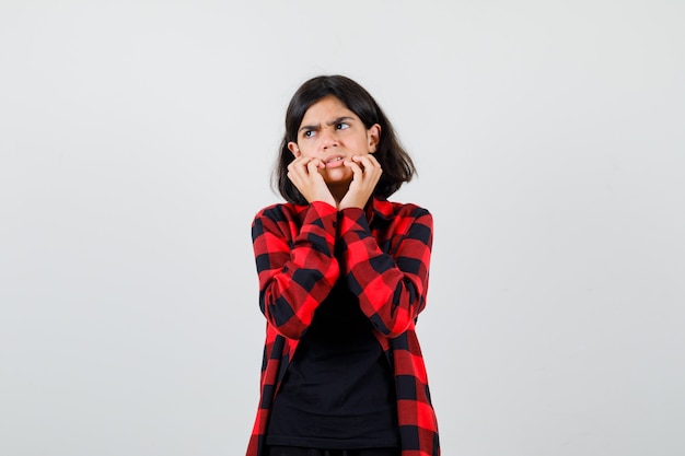 Chica adolescente en camiseta, camisa a cuadros cogidos de la mano cerca de la boca y mirando insatisfecho, vista frontal.