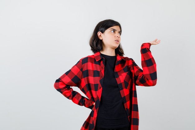 Chica adolescente en camiseta, camisa a cuadros fingiendo sostener algo y mirando con cuidado, vista frontal.