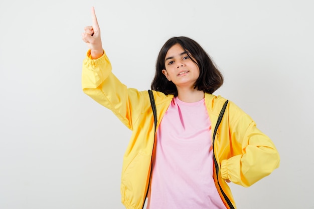 Chica adolescente en camiseta, chaqueta apuntando hacia arriba y mirando confiado, vista frontal.