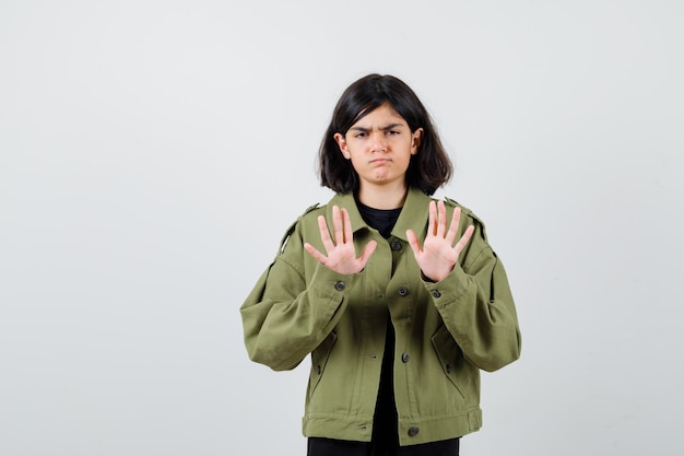 Chica adolescente en camiseta, chaqueta mostrando gesto de parada y mirando insatisfecho, vista frontal.