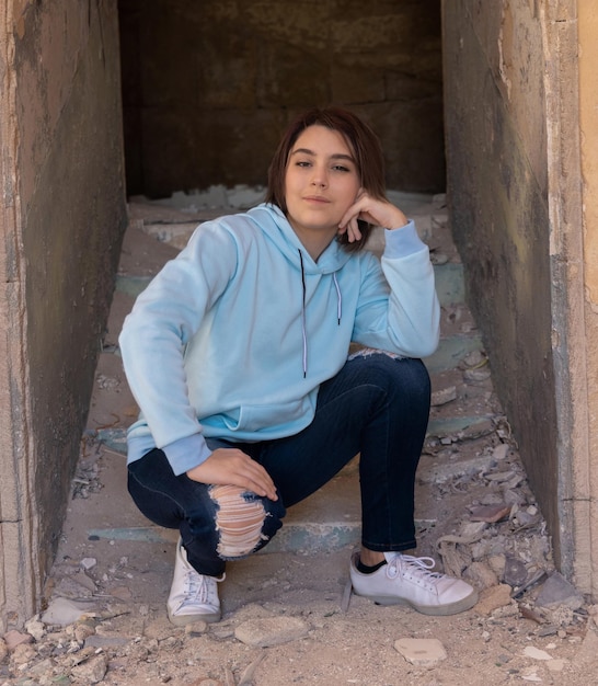 Chica adolescente con capucha azul claro sentada sobre sus ancas cerca de una pared