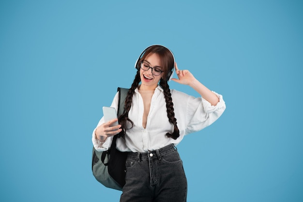 Chica adolescente caucásica feliz con coletas en gafas con auriculares estudiante con mochila usa teléfono