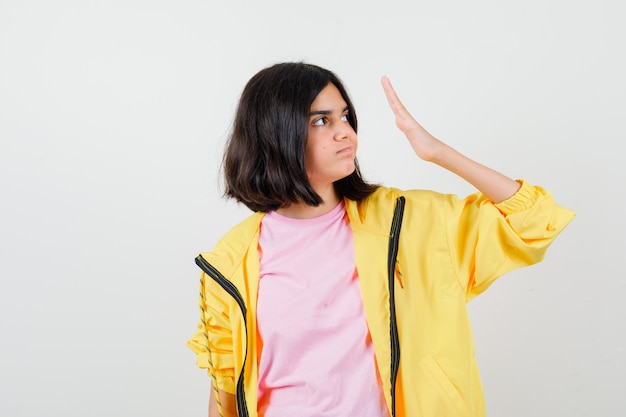 Chica adolescente en chándal amarillo, camiseta sosteniendo la palma cerca de la cara y mirando disgustado, vista frontal.