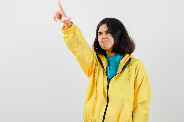 Foto chica adolescente en chaqueta amarilla apuntando hacia afuera y mirando disgustado, vista frontal.