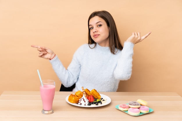 Chica adolescente comiendo gofres aislados en la pared beige apuntando con el dedo a los laterales y feliz