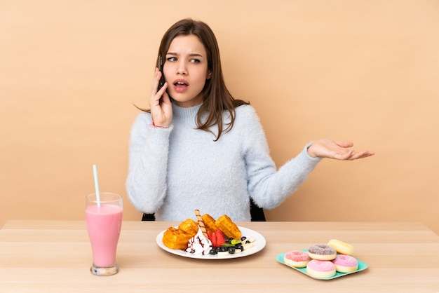 Chica adolescente comiendo gofres aislados en la pared beige manteniendo una conversación con el teléfono móvil con alguien