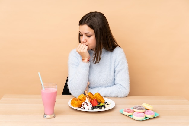 Chica adolescente comiendo gofres aislados en la pared de color beige con dudas