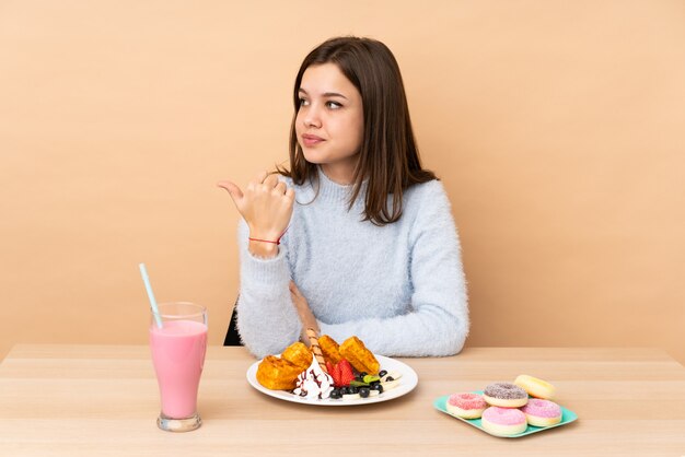Chica adolescente comiendo gofres aislados en la pared de color beige que apunta hacia un lado para presentar un producto