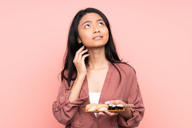 Chica adolescente comiendo sushi aislado en rosa pensando en una idea