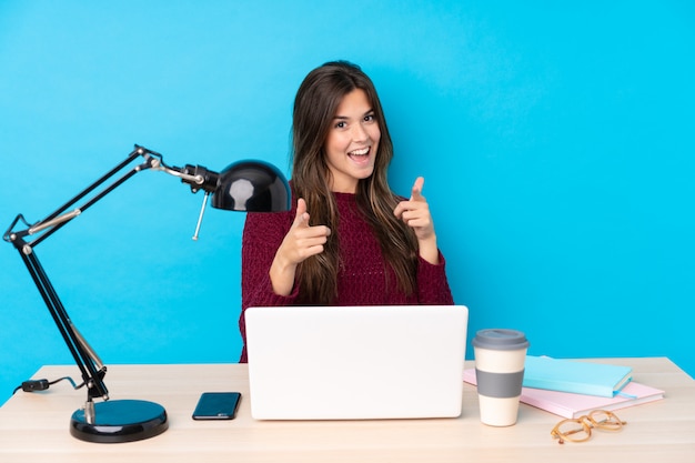 Chica adolescente con una computadora portátil en una mesa te señala con el dedo