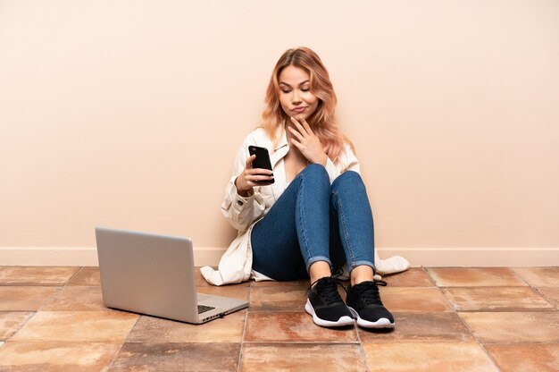 Chica adolescente con una computadora portátil sentada en el suelo en el interior pensando y enviando un mensaje