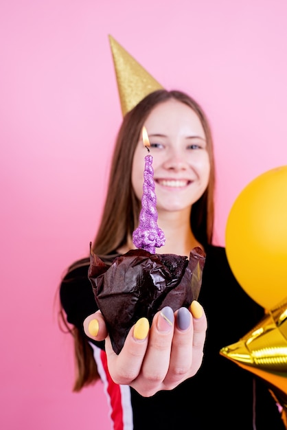 Chica adolescente en cumpleaños dorado sosteniendo pastel con vela, pidiendo un deseo sobre fondo de color rosa se centran en primer plano