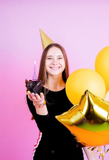 Chica adolescente en cumpleaños de oro sosteniendo muffin con velas, pidiendo un deseo sobre fondo de color rosa