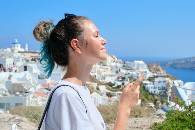 Chica adolescente descansando en la isla griega de Santorini, mujer mirando a otro lado, arquitectura de fondo blanco de la aldea de Oia, mar, cielo en las nubes, espacio de copia